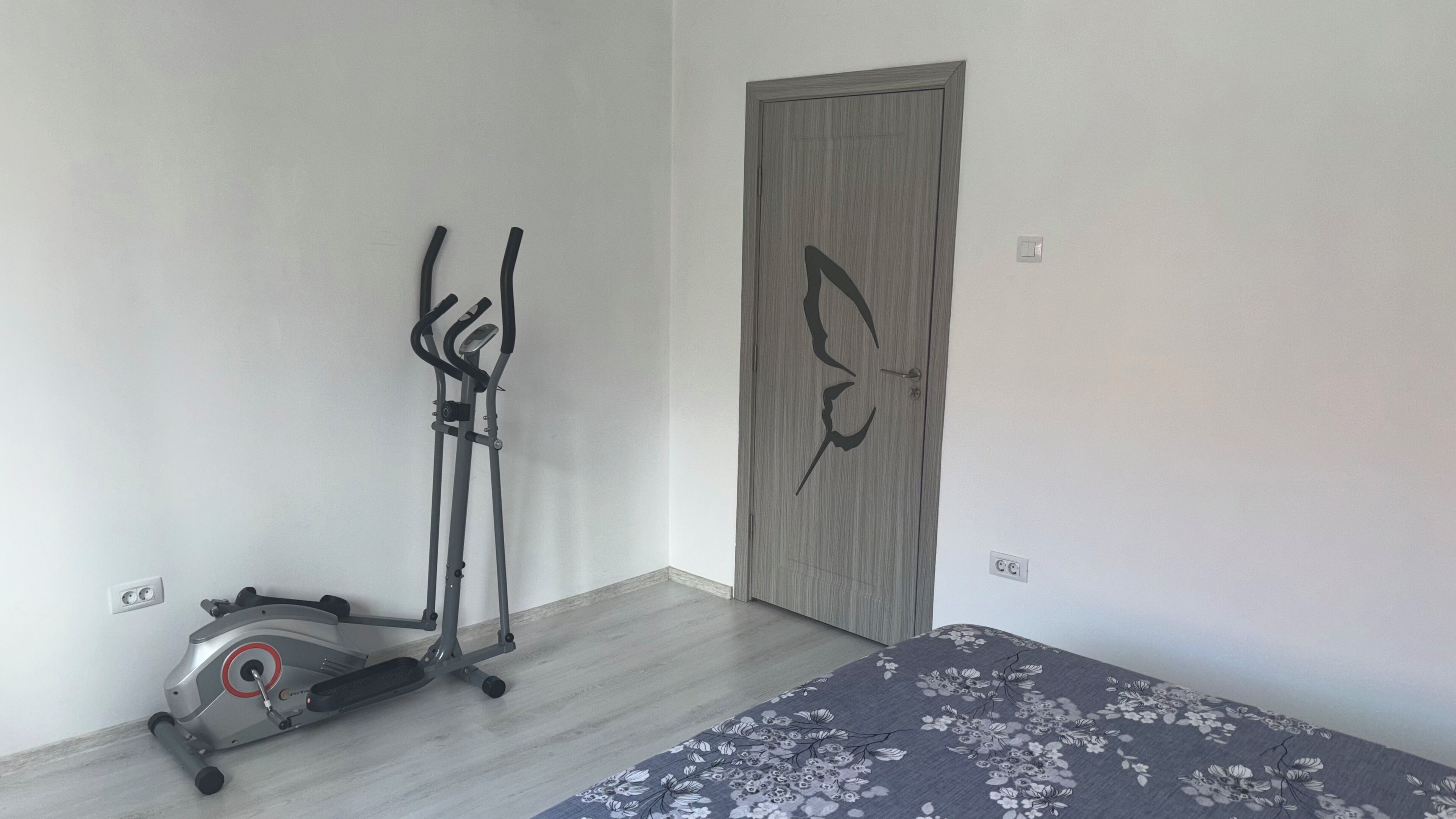 Een kamer met een hometrainer, een deur met een bloemistisch patroon en een bed met een bloemenbedekking.