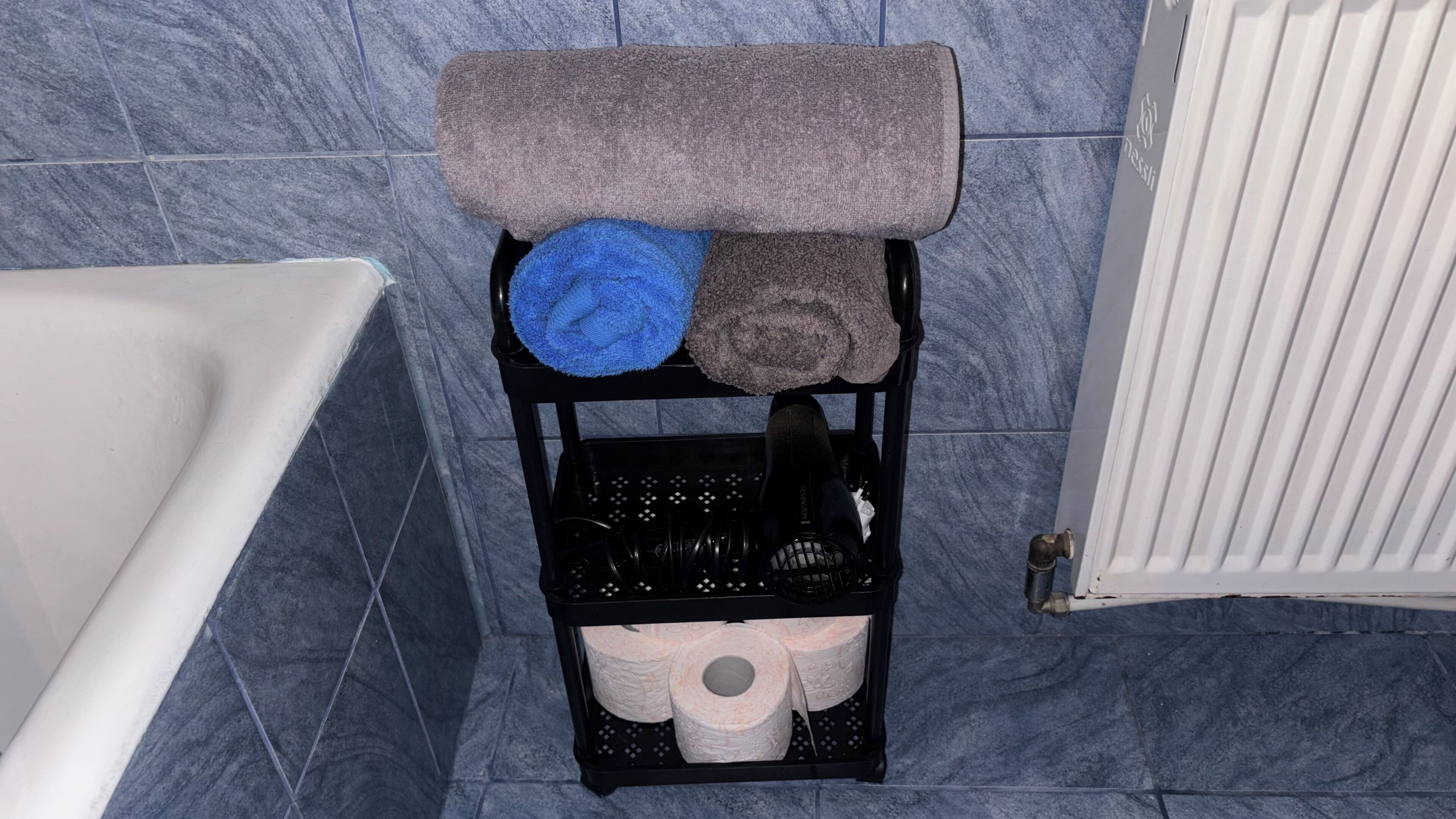 Casa de banho com banheira, prateleira preta com toalhas e papel higiénico, e radiador branco.
