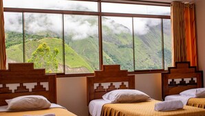 Quarto de hotel com três camas, uma janela grande com vista para as montanhas e cabeceiras de madeira.