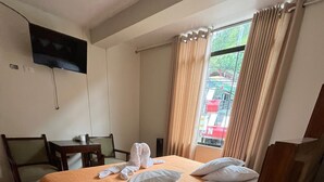 Quarto de hotel com cama, escrivaninha, cadeira, televisão e janela com cortinas.