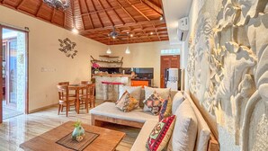 A spacious living area with a wooden ceiling, a stone fireplace, a dining set, and a kitchen in the background. - Janu Villa (Ubud)