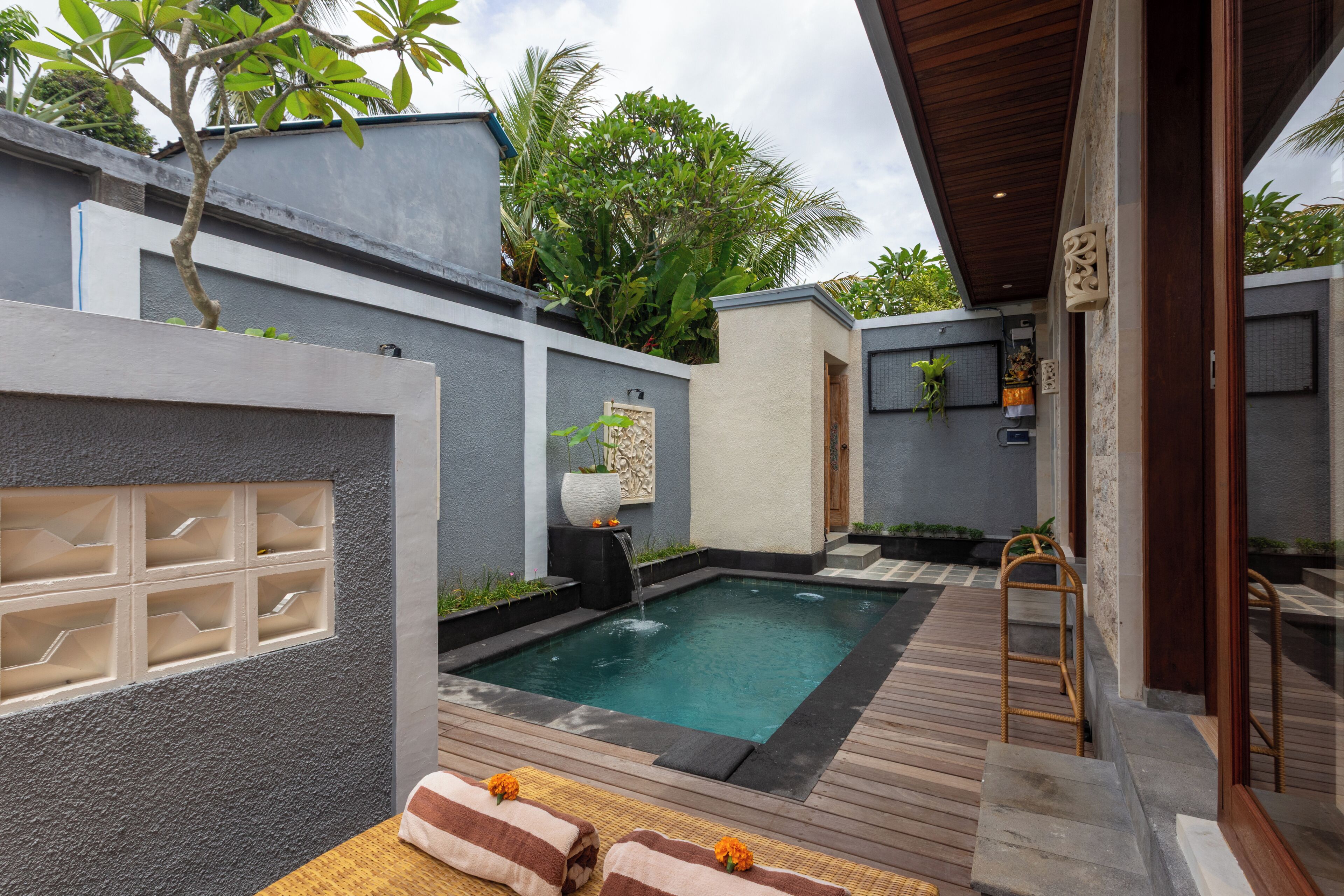 A modern outdoor pool area with a wooden deck, a small waterfall feature, and a lounge area with towels.