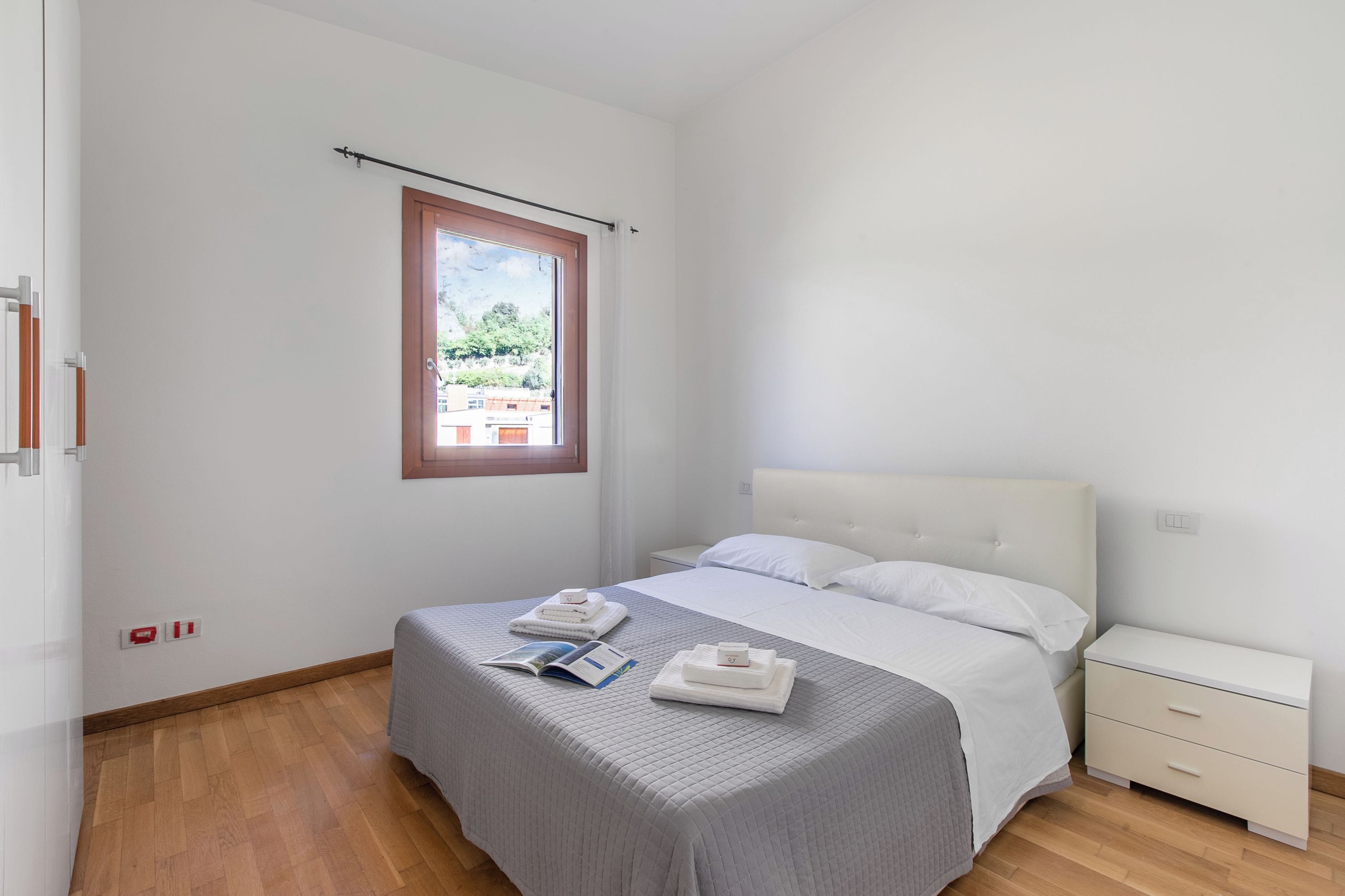 A bedroom with a bed, a nightstand, a window, and a view of buildings outside.