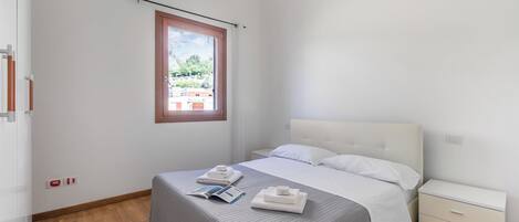 A bedroom with a bed, a nightstand, a window, and a view of buildings outside.