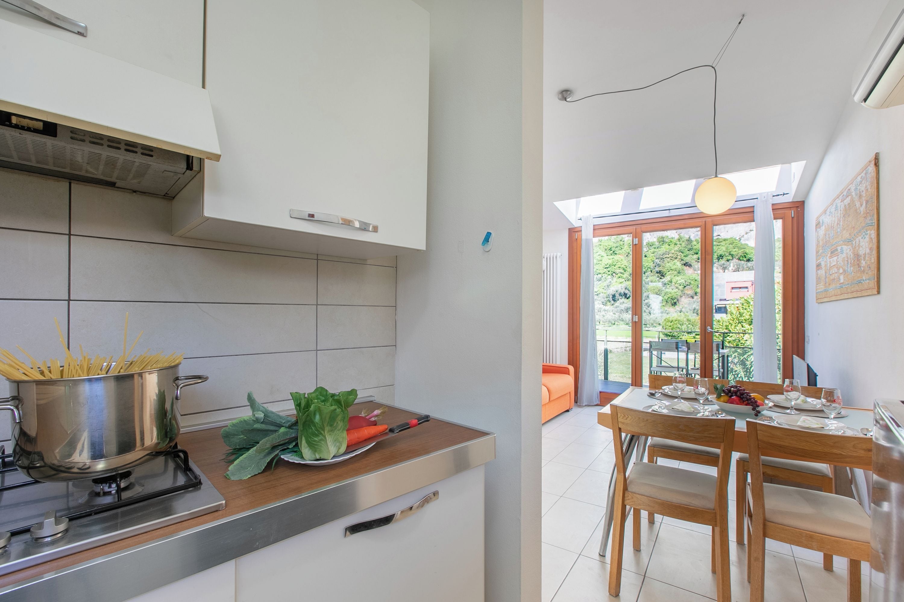 Una cocina con estufa, ollas y verduras sobre el mostrador, y una zona de comedor con una mesa preparada para dos.