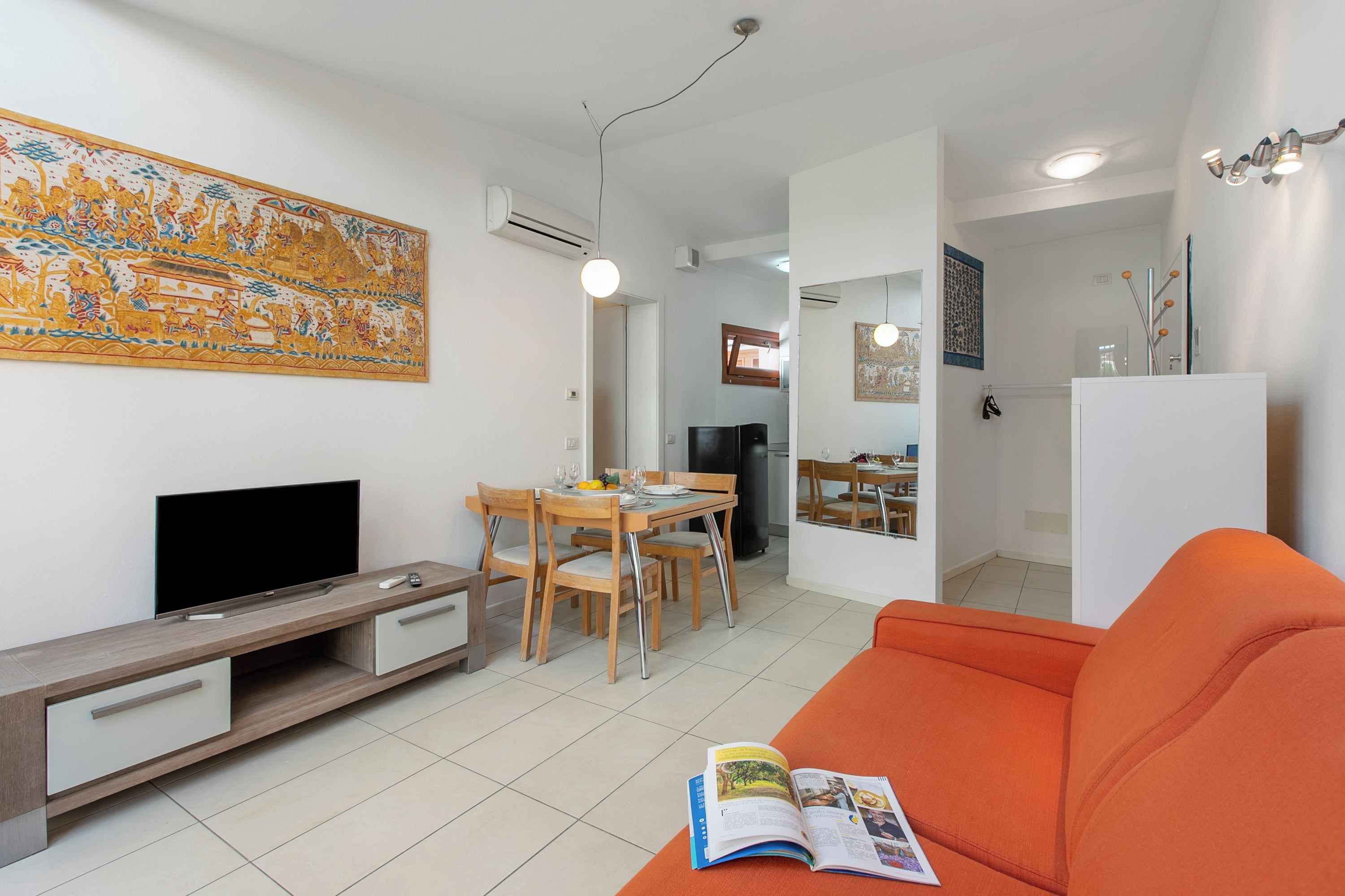 A modern living room with a flat-screen TV, a dining area with wooden chairs, a bright orange sofa, and a large decorative wall piece.