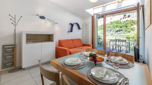 A dining area with a table set for four, a sofa, and a view of greenery outside. - Garda Therme Apartment With Balcony & Mountain Views (Arco)