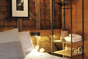A wooden cabin-style room with a bed, a shelf with towels, a framed picture on the wall, and a hanging rack.
