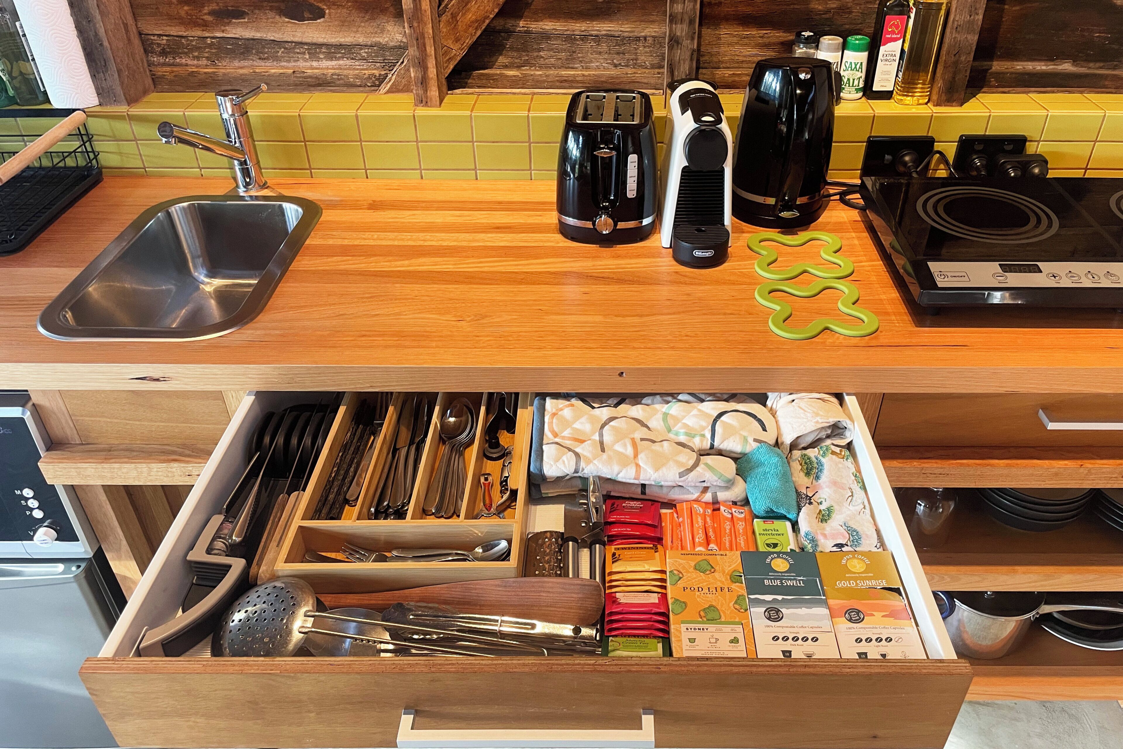 A kitchen with a wooden countertop, a sink, a toaster, a kettle, and a drawer with utensils and coffee pods.