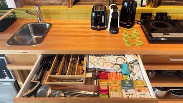 A kitchen with a wooden countertop, a sink, a toaster, a kettle, and a drawer with utensils and coffee pods.