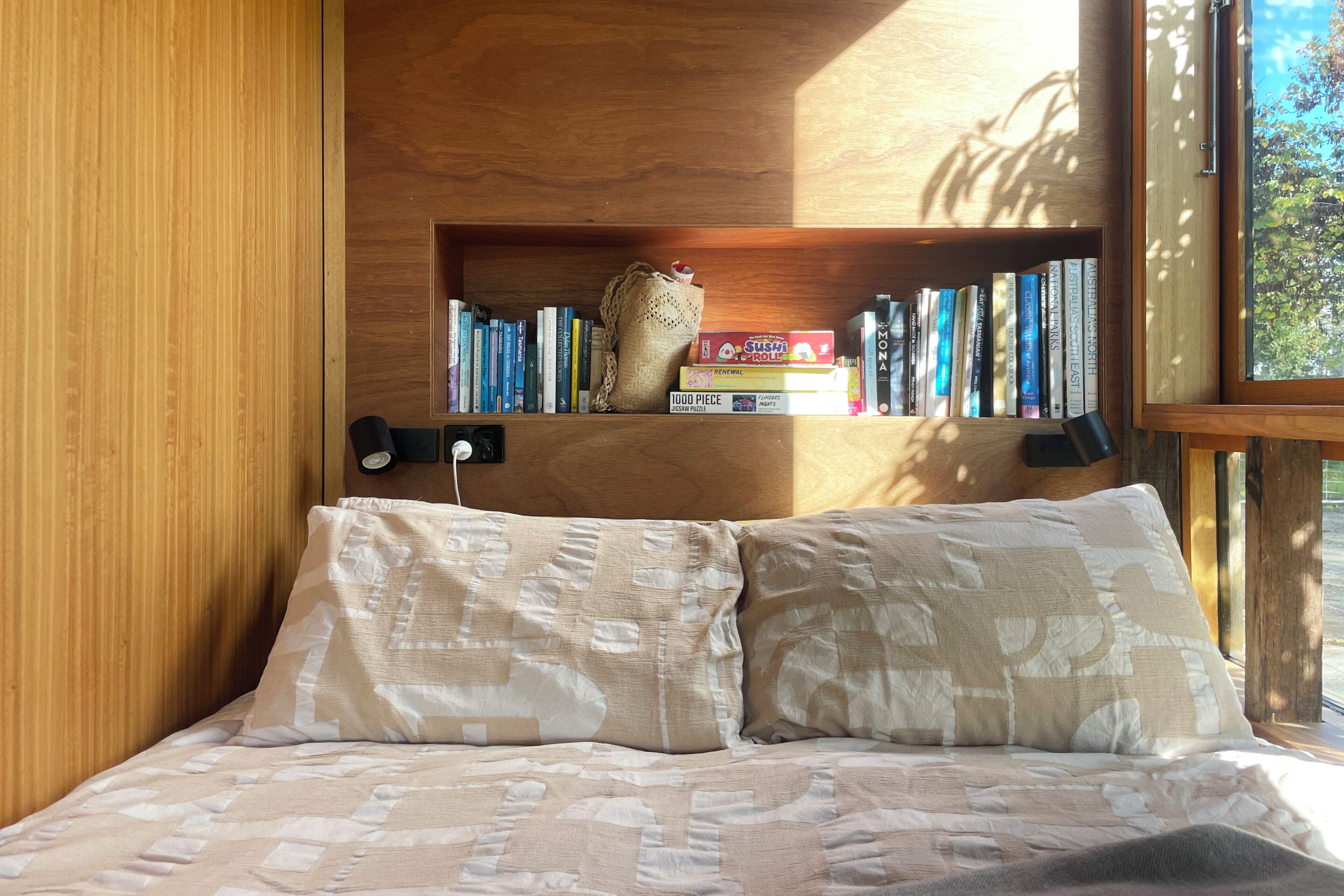 A bedroom with a wooden headboard, a bookshelf with books, a jigsaw puzzle, and a window with a view of trees.
