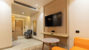 A modern hotel room with a flat-screen TV mounted on the wall, a small round table, two grey armchairs, an orange sofa, and a wooden reception desk. - The Archway Hotel (Haldwani)