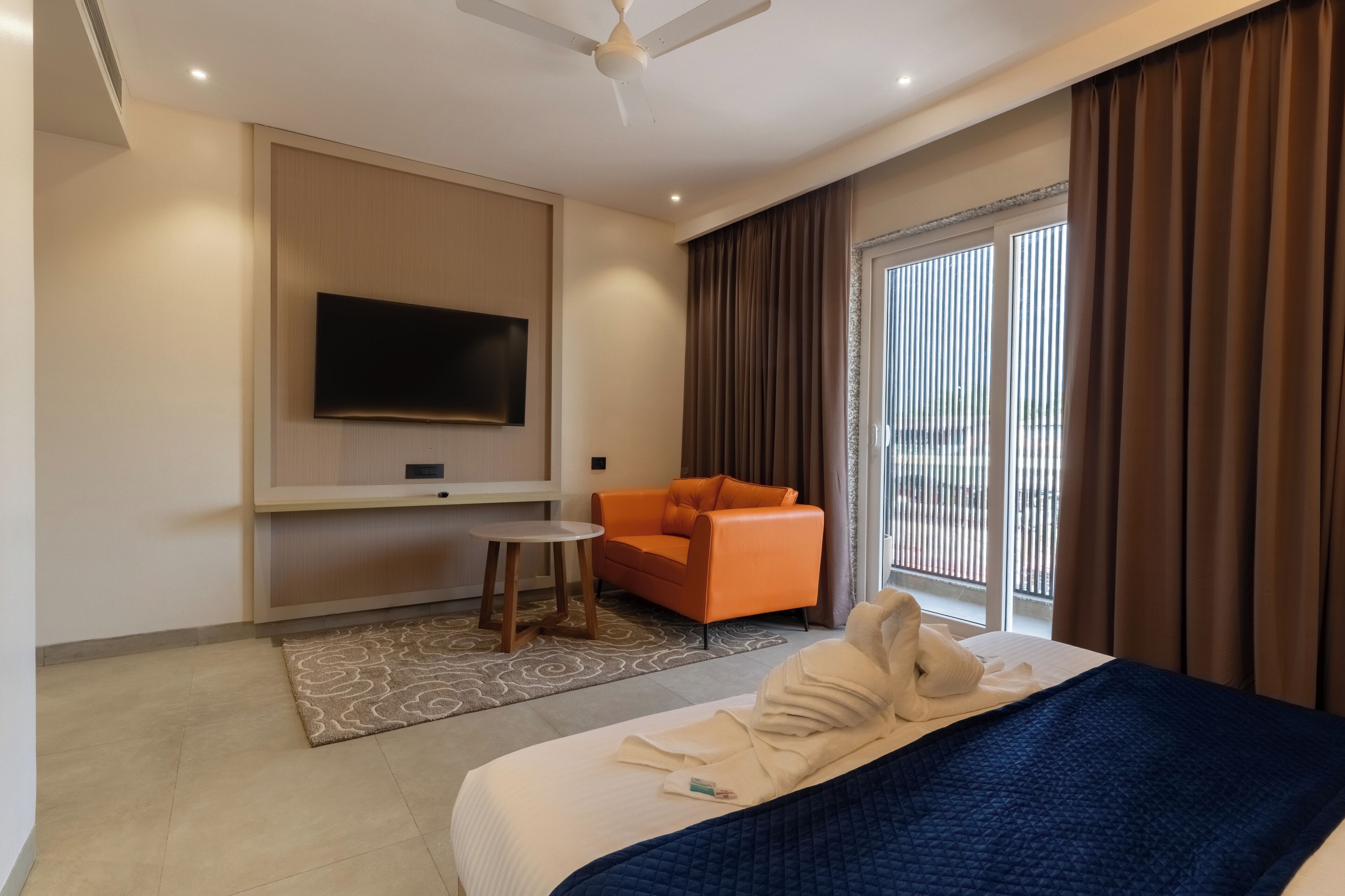 Habitación de hotel con cama, televisión montada en la pared, una silla naranja y un ventanal con cortinas.
