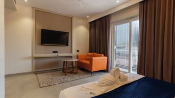 Habitación de hotel con cama, televisión montada en la pared, una silla naranja y un ventanal con cortinas.