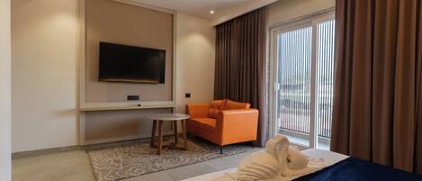 Habitación de hotel con cama, televisión montada en la pared, una silla naranja y un ventanal con cortinas.