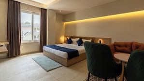 A hotel room with a bed, a desk, a chair, a small table, and a view of the outside. - The Archway Hotel (Haldwani)
