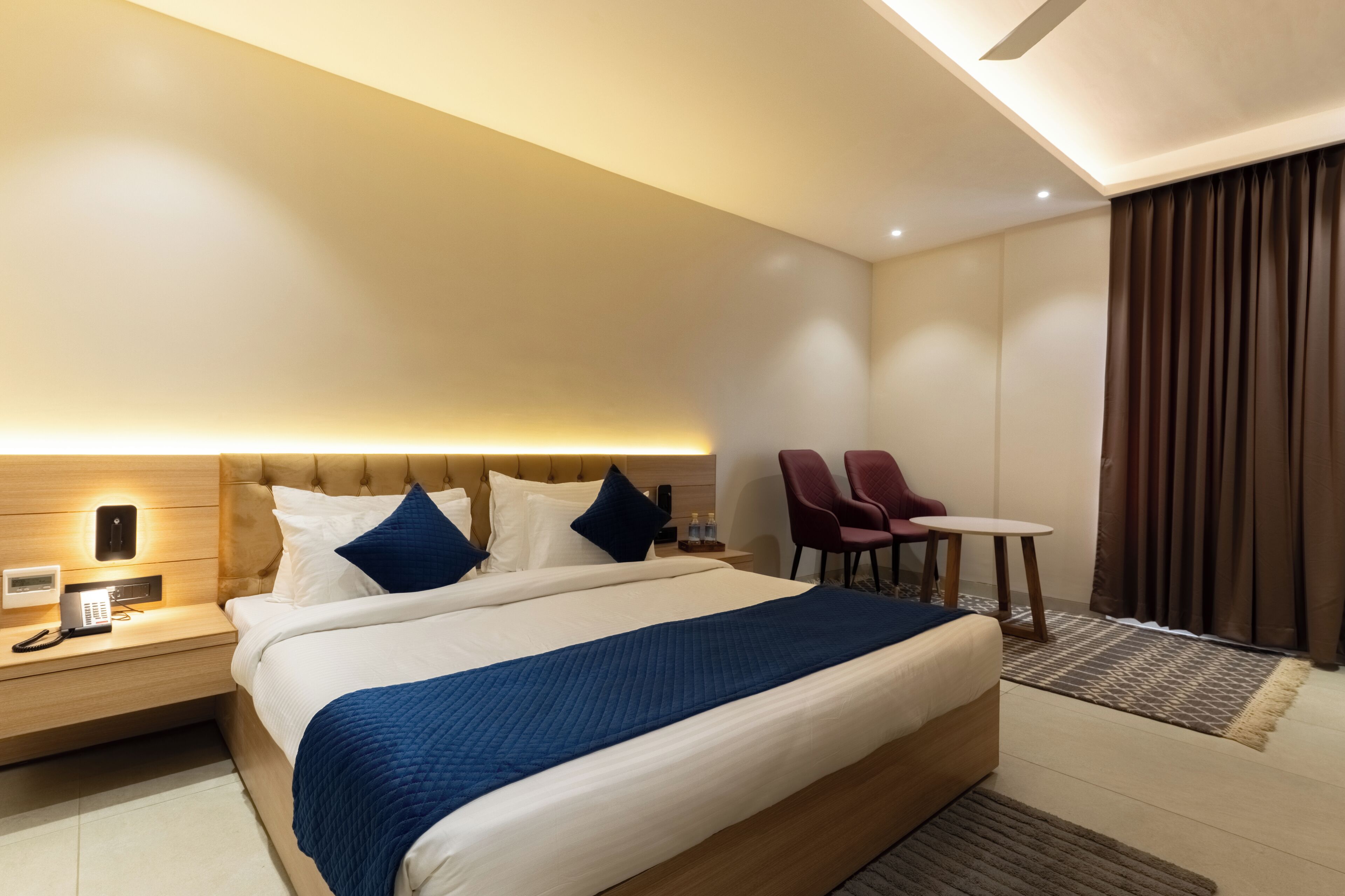 A modern hotel room with a large bed, bedside table with a phone, a small table with chairs, and a ceiling fan.