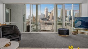 A modern living room with a large window, a black leather sofa, a round coffee table, and a flat-screen TV. - Exclusive Stays - Southgate (Southbank)