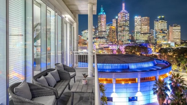 A balcony with wicker furniture, a view of a city skyline at night, and a lit building with a circular roof. - Exclusive Stays - Southgate (Southbank)