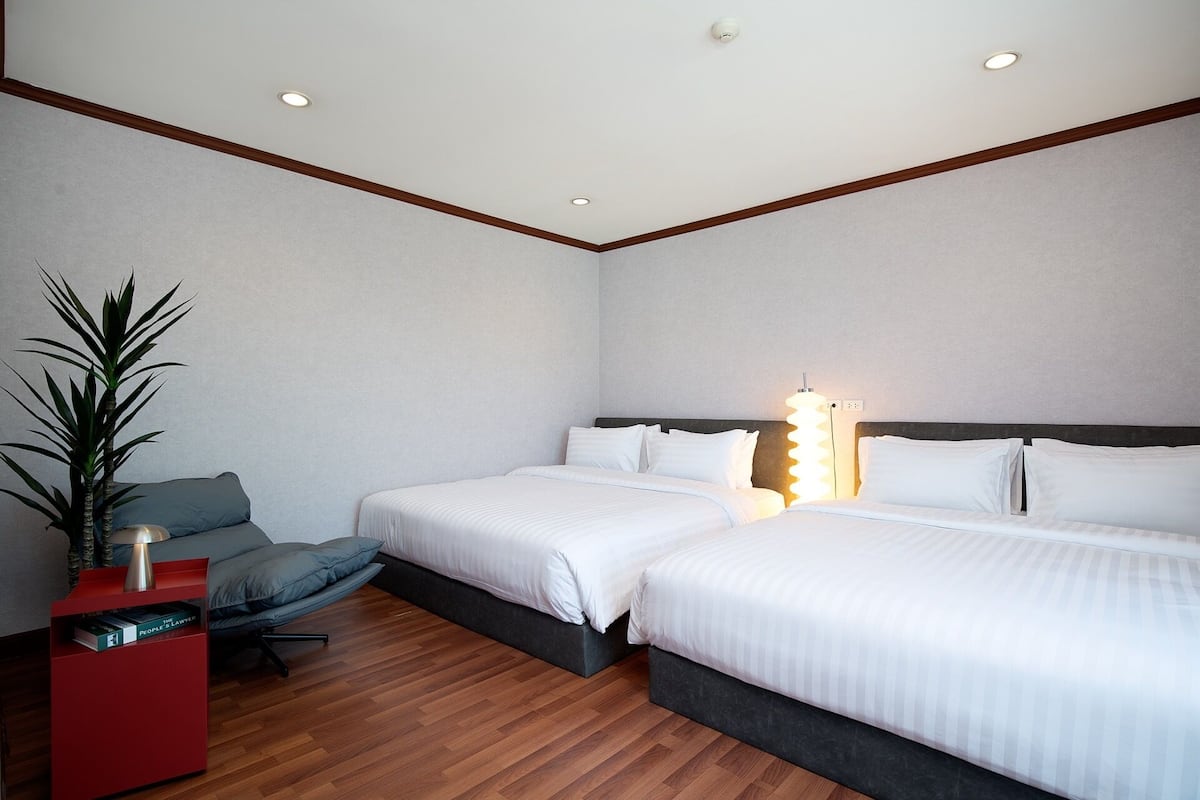 A hotel room with two beds, a nightstand, a chair, a potted plant, and a wall-mounted light fixture.
