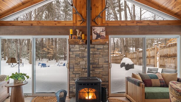 L’intérieur chaleureux d’un chalet avec une cheminée en pierre, des meubles en bois et de grandes fenêtres donnant sur une forêt enneigée.