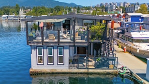 Une péniche aménagée en maison flottante, dotée d’une terrasse, de sièges extérieurs et offrant une vue sur l’eau et la ville en arrière-plan.