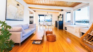 Un salon moderne avec un plafond en bois, un canapé, des poufs, un coin repas et de grandes fenêtres offrant une vue dégagée.