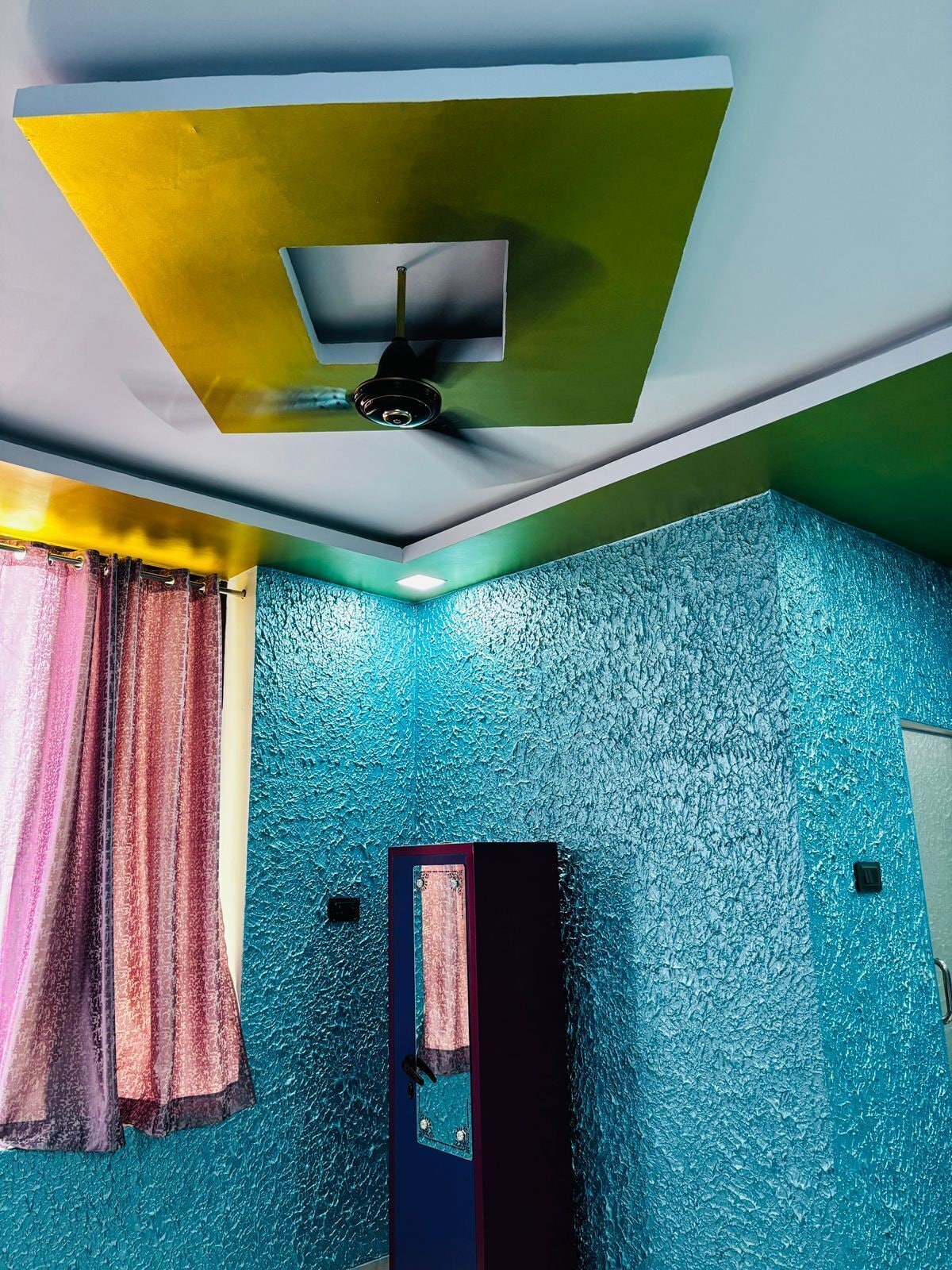 Quarto com parede azul texturizada, ventilador de teto e janela com cortinas.