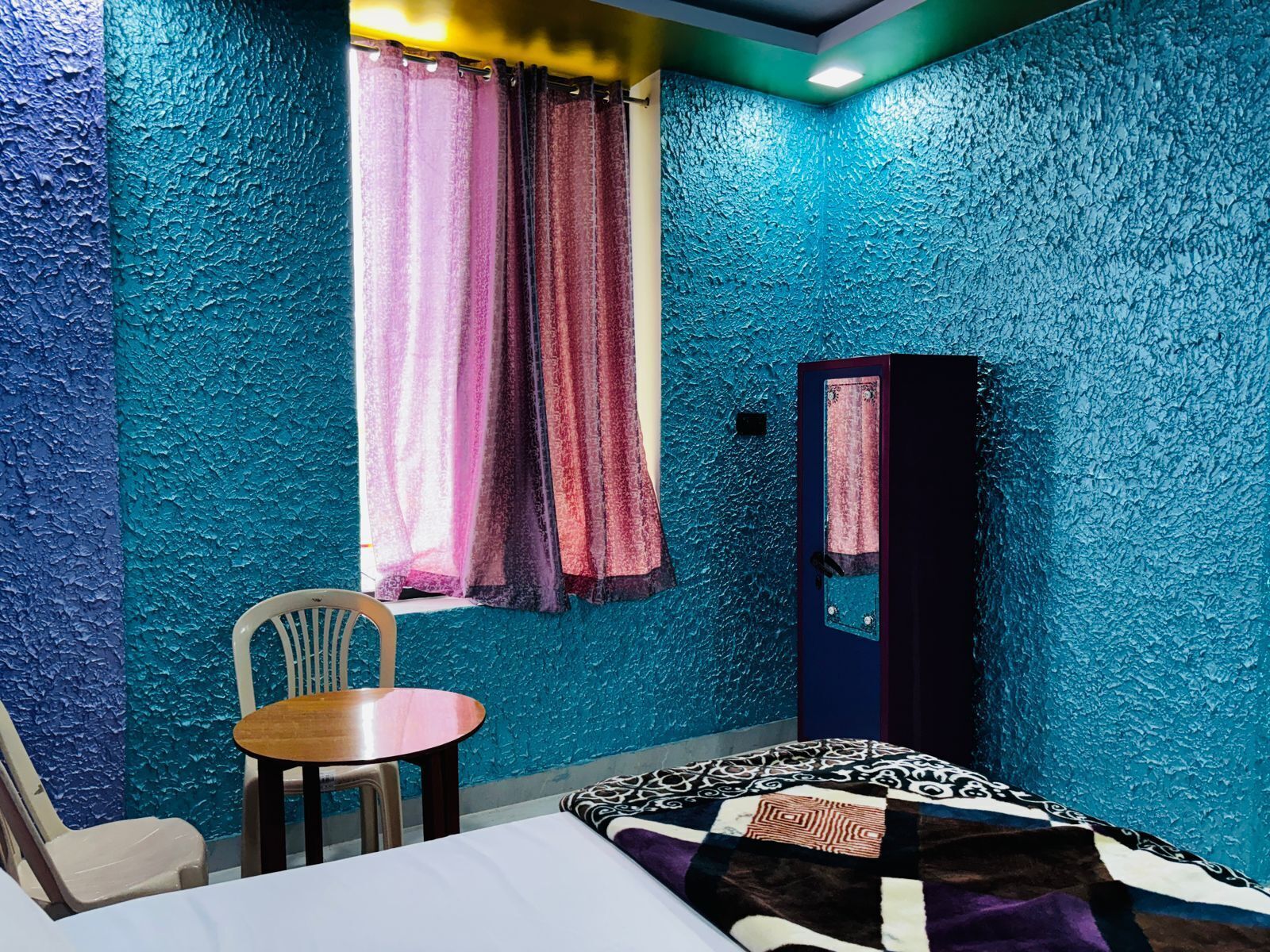 Una habitación con cama, una silla, una mesita, una ventana con cortinas y un armario.