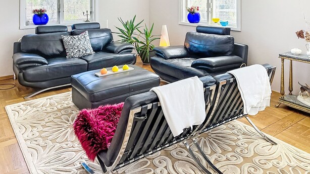 Una sala de estar moderna con dos sofás de cuero negro, una mesa de centro con fruta, un tapete estampado y un ventanal con persianas.