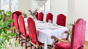 Comedor con una larga mesa dispuesta para seis comensales, sillas tapizadas en rojo, un candelabro y vistas a través de una ventana con persianas.