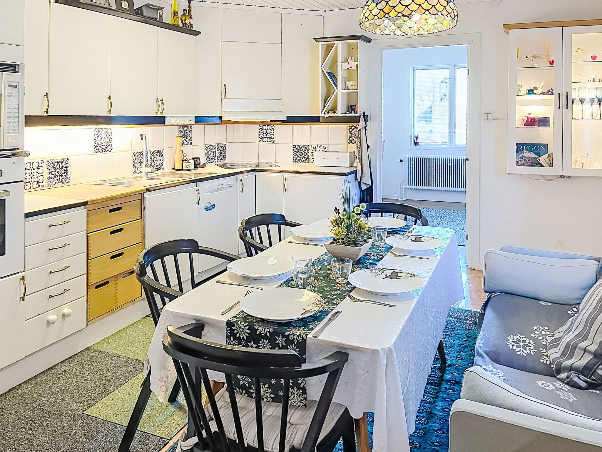 A modern kitchen with white cabinets, a dining area with a long table set for four, and a blue patterned rug.