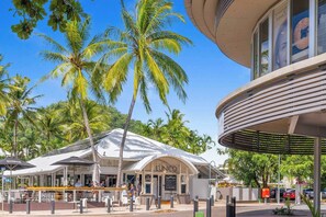 A restaurant with outdoor seating, palm trees, and a modern building with a curved facade. - Oceanfront Gem at Trinity Beach - Pool - Sea View (Trinity Beach)