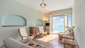 Ein Zimmer mit einer Couch, zwei Schaukelstühlen, einem kleinen Tisch mit einer Vase und Blumen sowie Blick auf das Meer durch eine offene Tür.