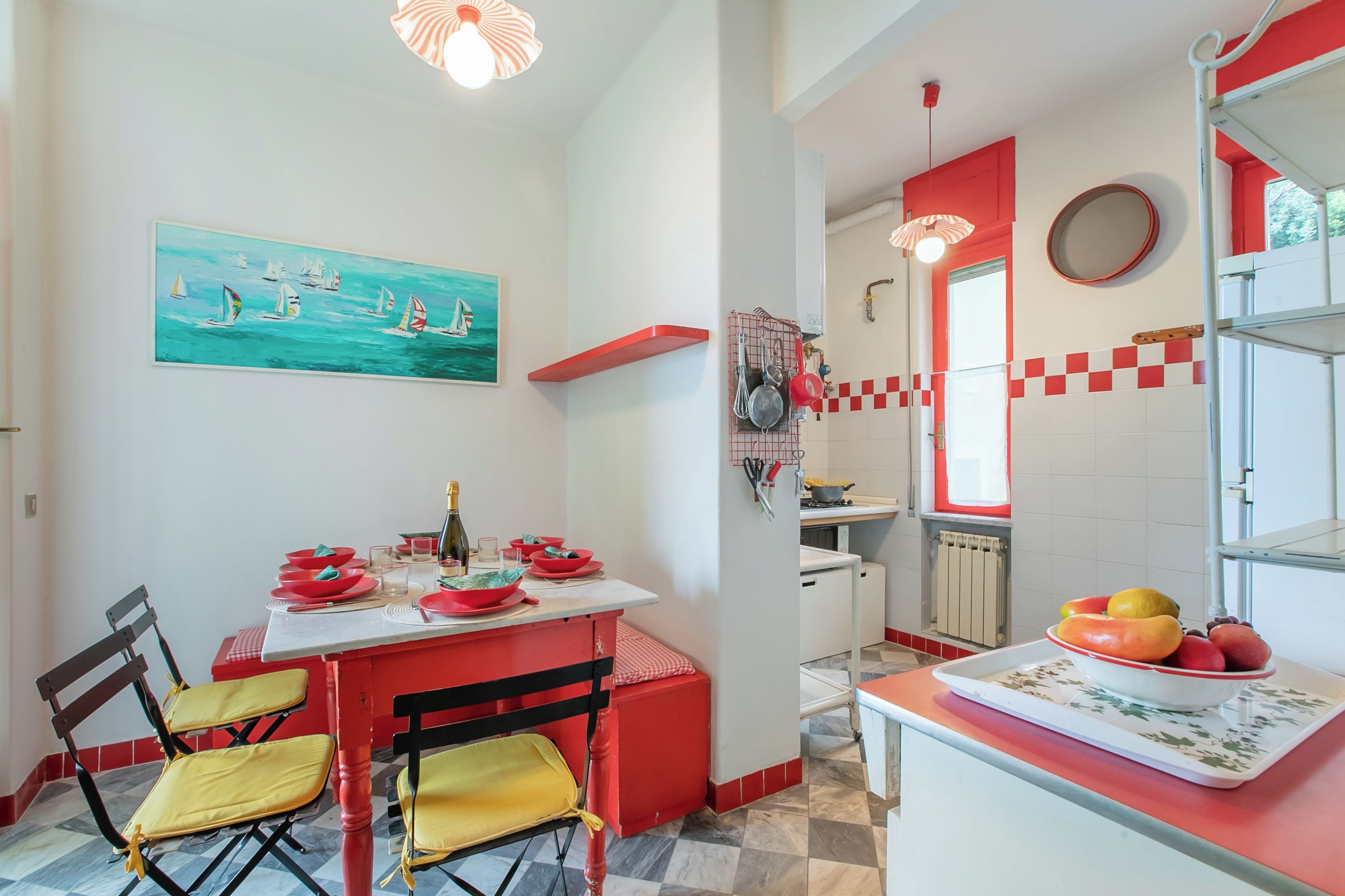 Cozinha com mesa de jantar posta para quatro pessoas, tigela de frutas e pintura de veleiros.