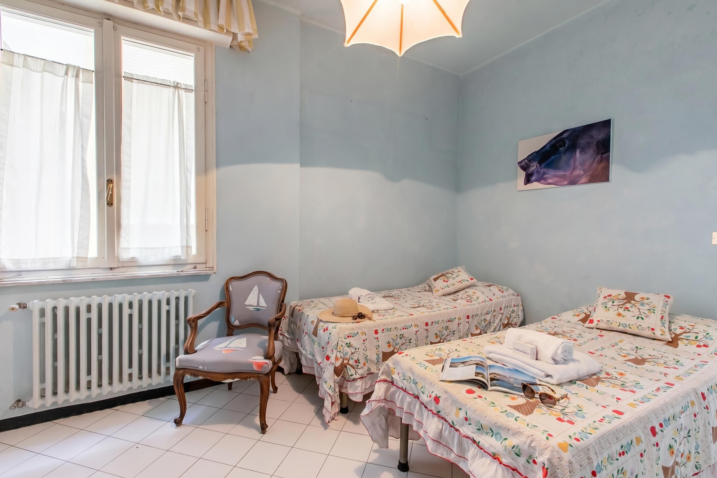 A room with two beds, a chair, a radiator, and a window with curtains.