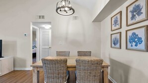 A dining area with a wooden table and wicker chairs, a chandelier, and framed artwork on the wall. - Gulf Shores Condo with Pool & Hot Tub (Gulf Shores)