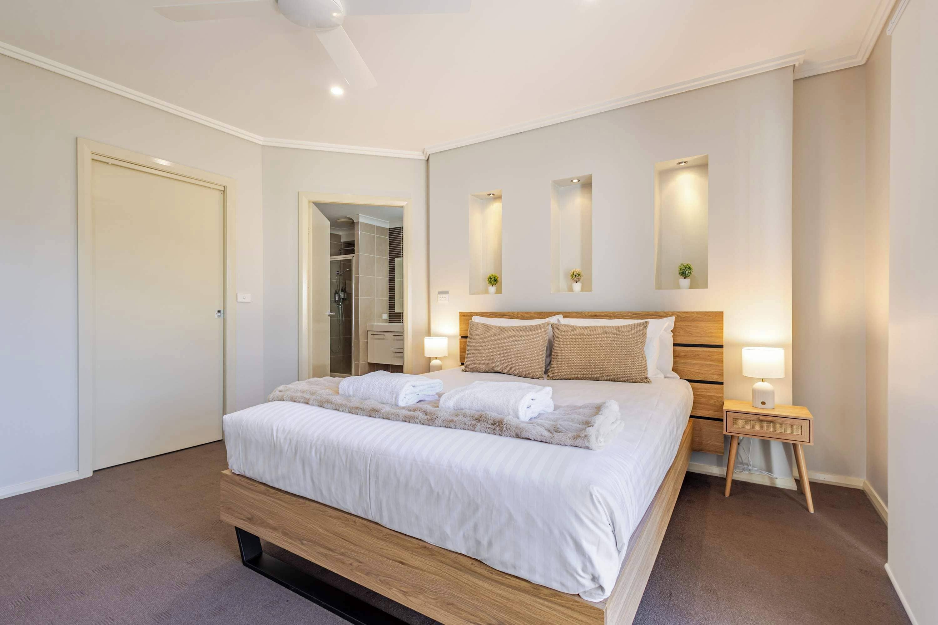 A modern hotel room with a large bed, bedside lamps, a wooden headboard, and a shower area visible through an open door.