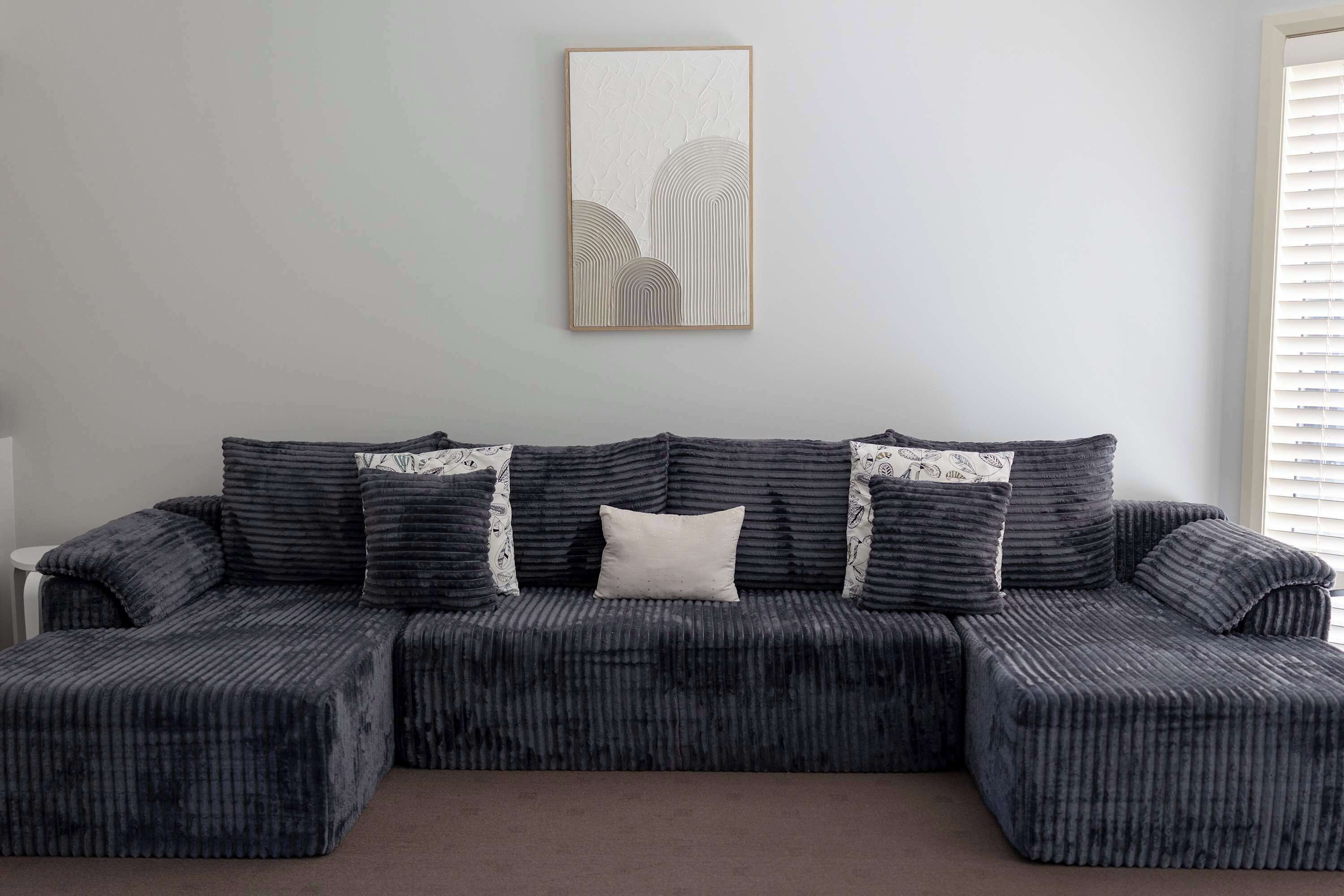 Ein modulares Sofa mit strukturierter Stoffbezüge und gemusterten Kissen, darüber ein gerahmtes Kunstwerk und rechts daneben ein Fenster mit Jalousien.