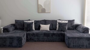 Een modulaire bank met een geruite bekleding en decoratieve kussens, een lijstwerk erboven en een raam met zonwering aan de rechterkant.