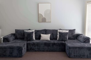 A sectional sofa with a textured fabric and patterned cushions, a framed artwork above it, and a window with blinds on the right.