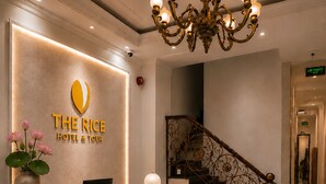 A hotel reception desk with a modern design, a chandelier, a staircase, and a sign indicating the exit. - The Rice Hotel & Tour - 27 Nguyen Thai Binh (Ho Chi Minh City)