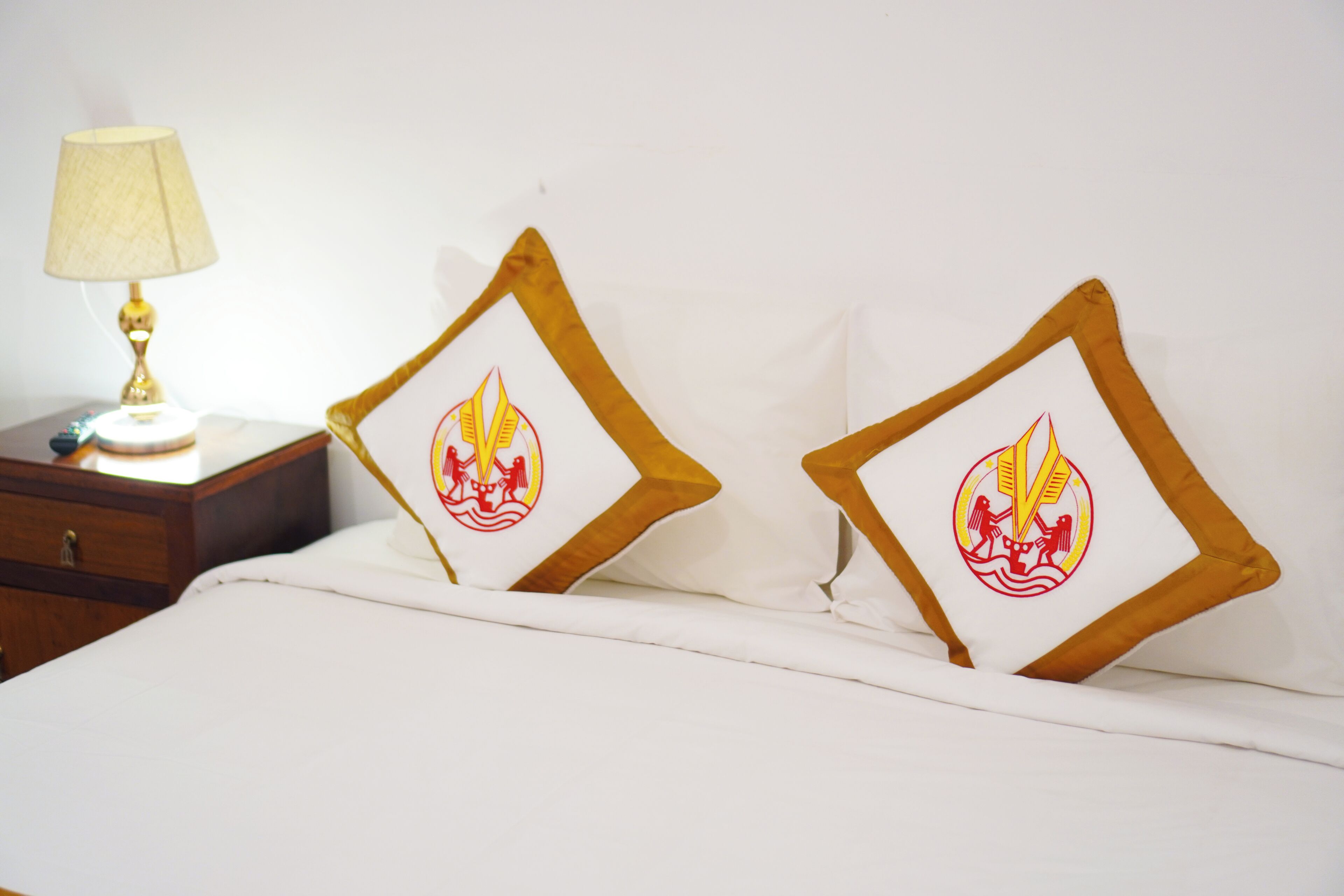Una cama con dos almohadas que presentan un emblema rojo y amarillo, una mesita de noche con una lámpara y una mesita auxiliar de madera.