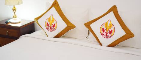 Una cama con dos almohadas que presentan un emblema rojo y amarillo, una mesita de noche con una lámpara y una mesita auxiliar de madera.