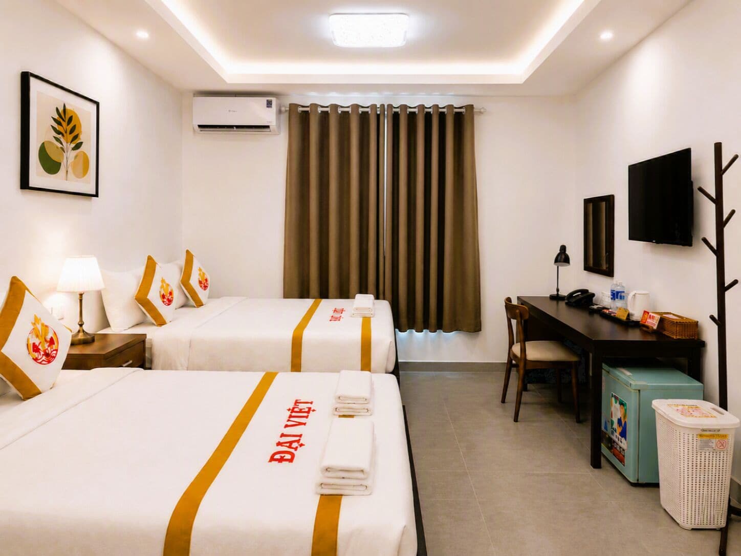 A hotel room with two beds, a desk, a chair, a TV, an air conditioner, and a trash bin.