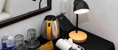 Ein Hotelzimmer mit einem schwarzen Schreibtisch, einer Kaffeemaschine, einem Telefon, einer Lampe, einem Spiegel und einem Tablett mit Wasserflaschen und Snacks.