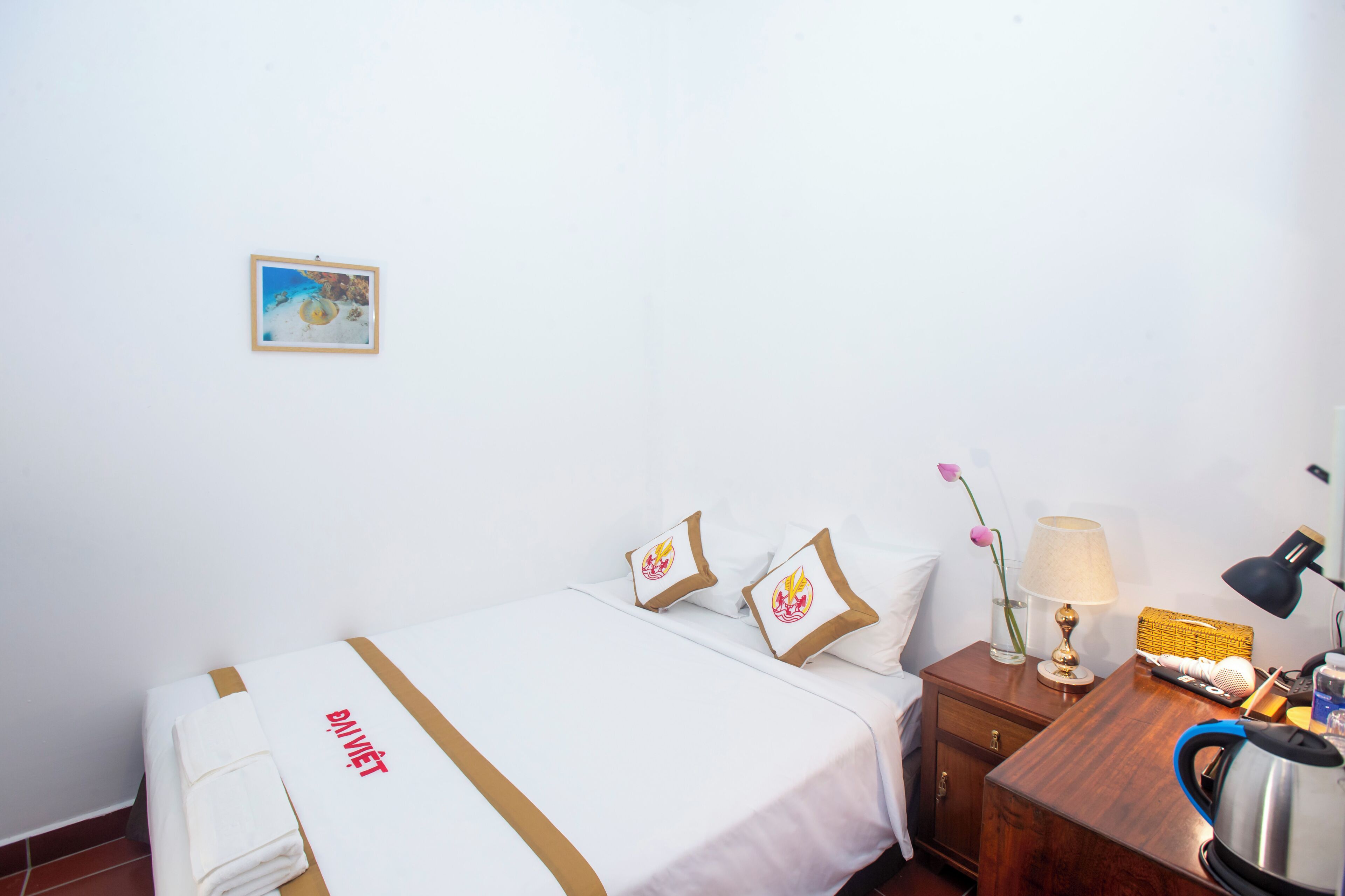 A hotel room with a bed, pillows, a nightstand, a lamp, a kettle, and a framed picture on the wall.