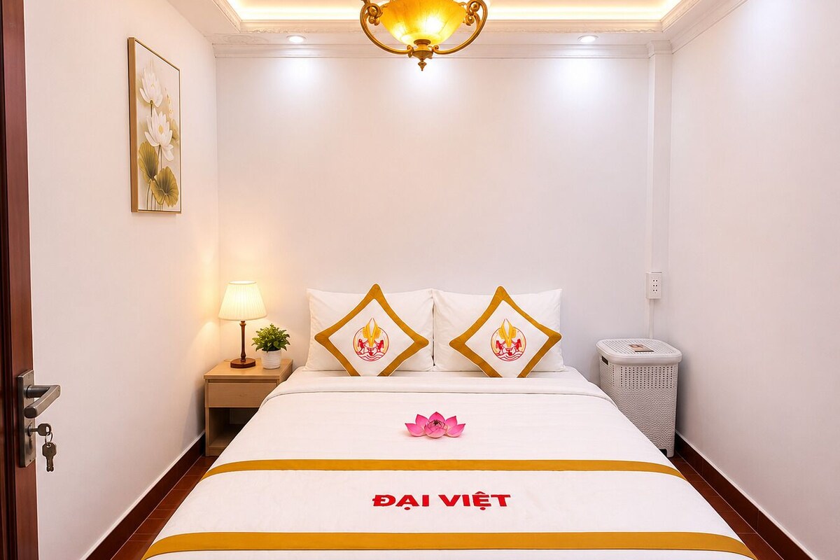 A hotel room with a bed, a nightstand with a lamp, a wall-mounted picture, and a trash bin.