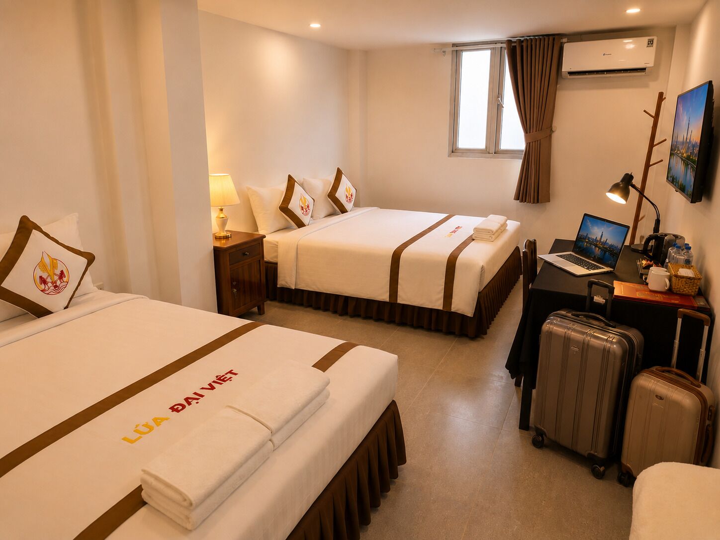 A hotel room with two beds, a desk with a laptop, a suitcase, and an air conditioning unit.