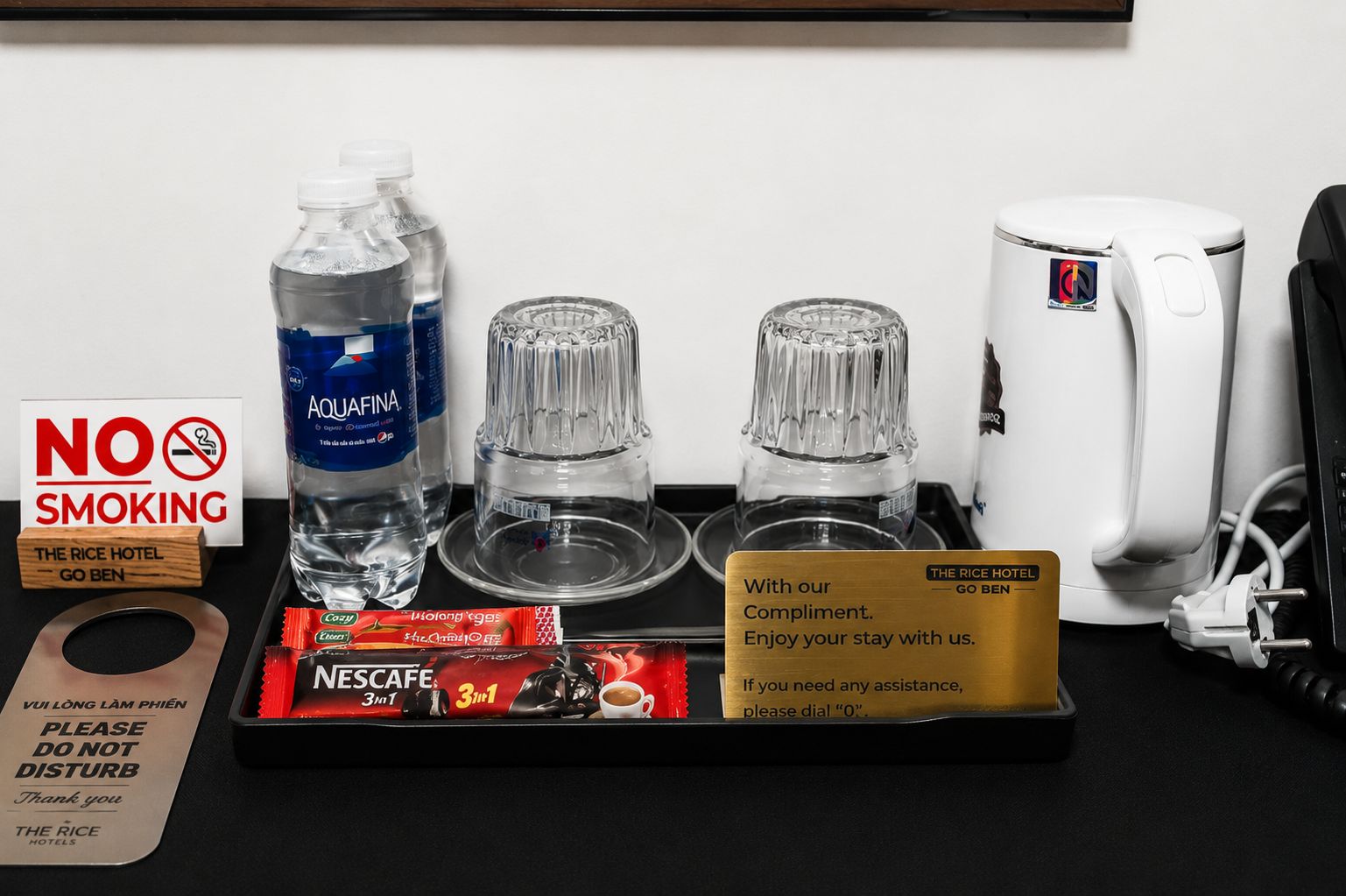 A hotel minibar setup with a water bottle, two glasses, a coffee maker, and a tray with a Nescafe coffee pack and a complimentary card.